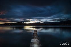 As the clouds slide overhead the small wooden pier reaches out over the waters of Lake Hayes into the last light of the day. Lake Hayes, Otago, South Island, New Zealand - landscape photography