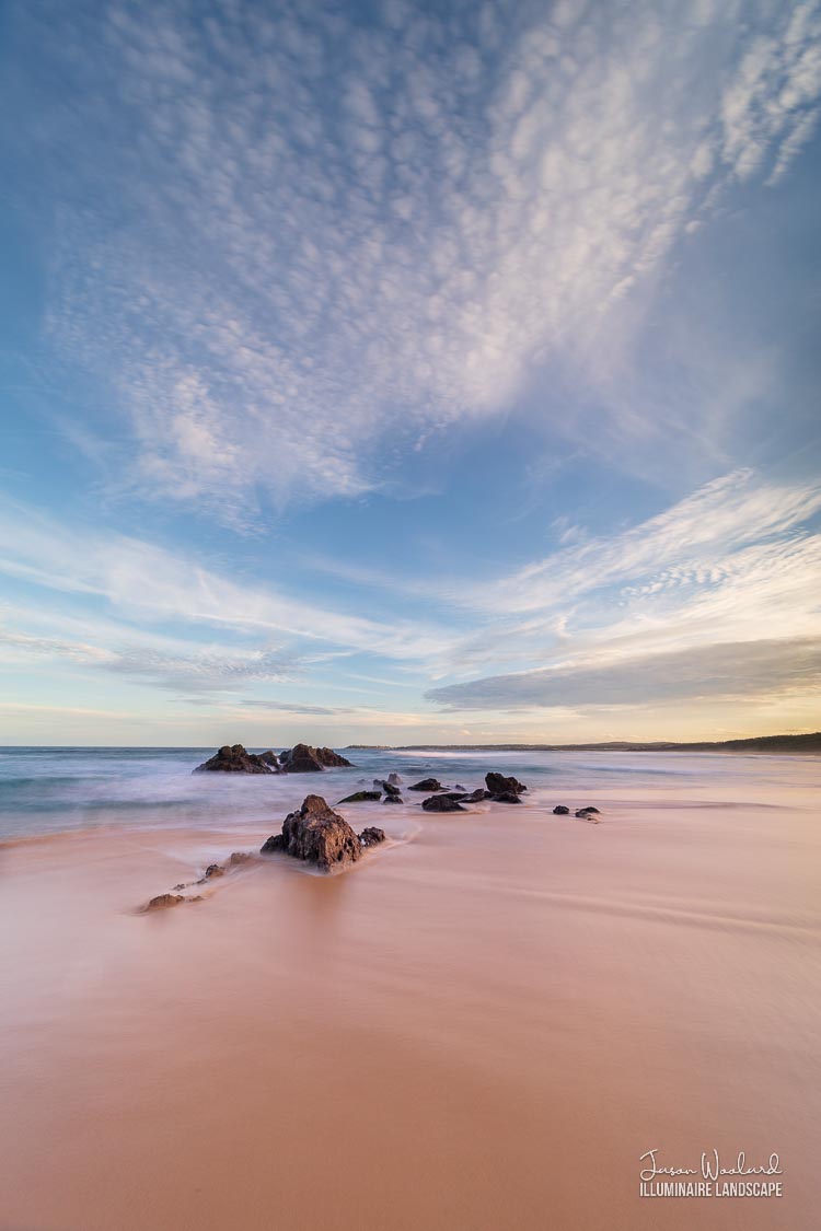 Soft speckled clouds tower over sea swirled rocks sitting in the sand. Haywards Beach, Wallaga Lake, New South Wales